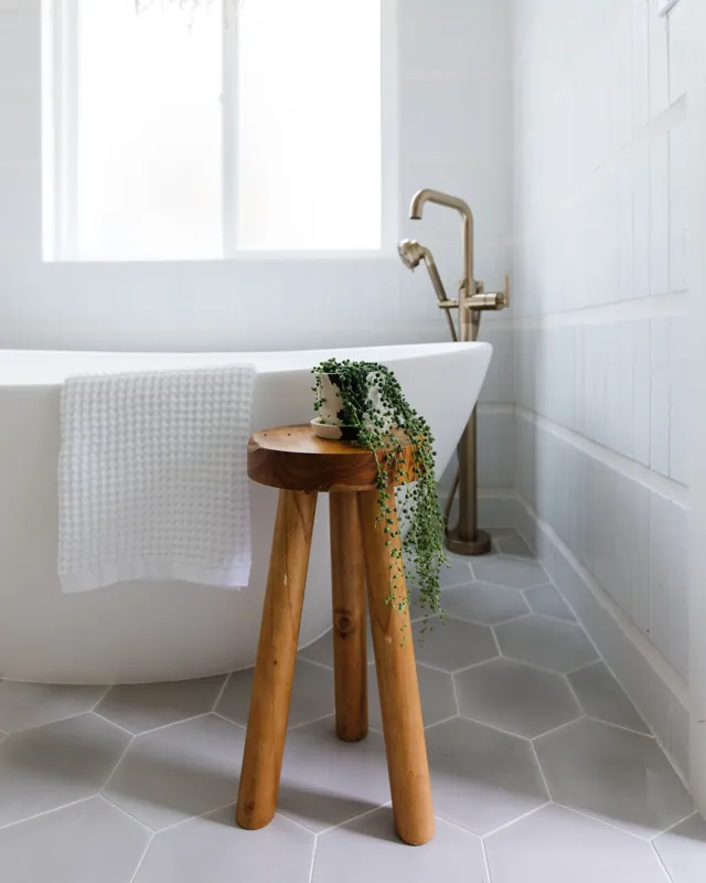 A bathroom with a white bathtub, a wooden stool with a potted green plant, and a window in the background.