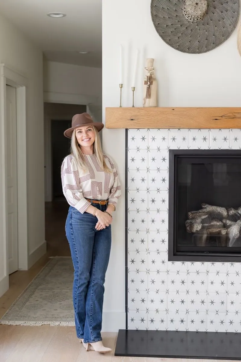 A woman with blonde hair, wearing a beige hat, beige patterned blouse, and blue jeans, standing next to a fireplace with white patterned tiles and a wooden mantel.