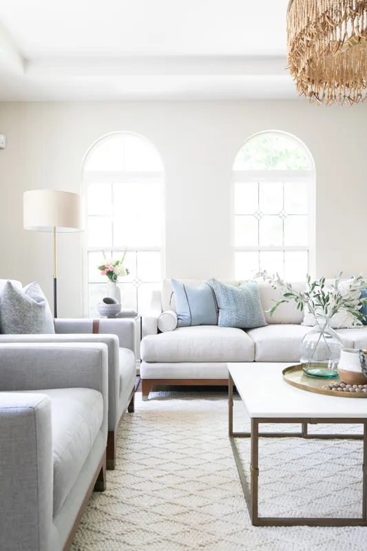 Bright living room with two large arched windows, white walls, and light-colored seating. Includes a beige sofa with blue pillows, a matching armchair, a white coffee table with a glass vase of greenery, and a tall floor lamp with a white shade.