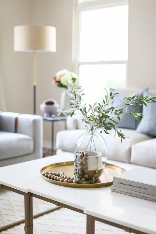 A living room with white furniture, a gold tray on a white coffee table holding a small white and brown pot with a beaded necklace, a glass vase with green leaves, and a book titled 'The Museum of Modern Art, New York'.