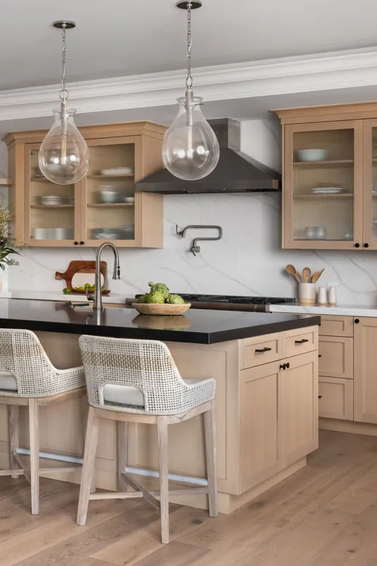 Modern kitchen with light wood cabinets, black countertop, marble backsplash, and two glass pendant lights over the island. There are two wicker chairs at the island.