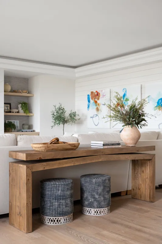 A living room features a wooden table with a large clay vase filled with greenery and a stack of books, two black patterned stools underneath, a cream-colored sofa, abstract paintings on the wall, and shelving with decor items.
