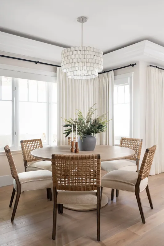 A dining room with a round wooden table, six woven and upholstered chairs, a large gray vase with greenery, two candlesticks, and natural light from large windows with white curtains. A modern white pendant light hangs from the ceiling.