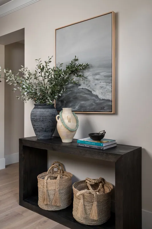 Decorative hallway table with vases, books, and baskets in front of a wall with a large framed landscape painting.