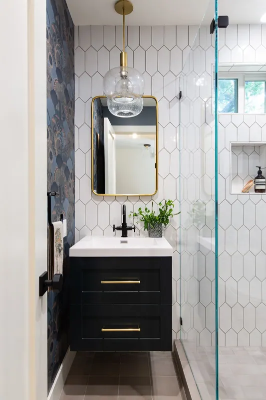 Modern bathroom with a black vanity, a white sink, a black faucet, a mirror with a gold frame, and a glass pendant light. A small potted plant and a towel sit on a black towel ring, and the wall behind the vanity has blue patterned wallpaper.