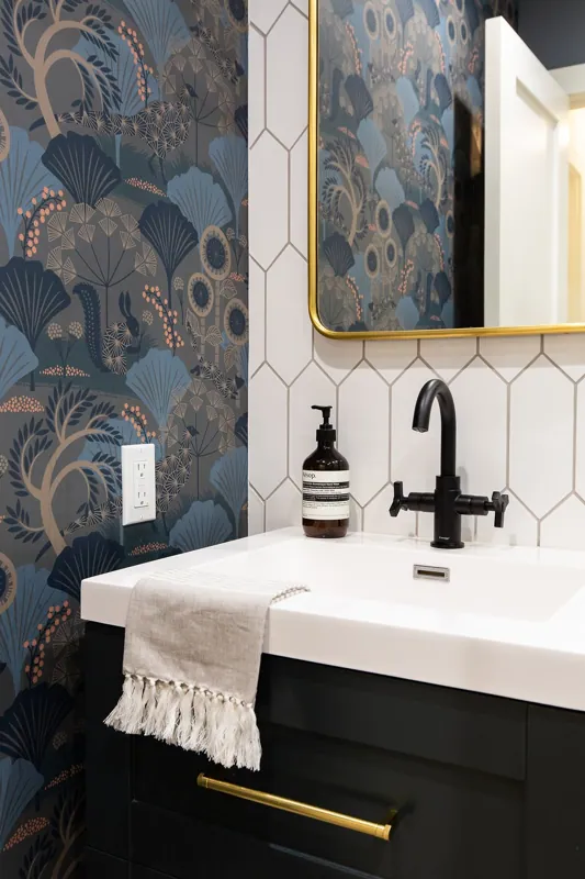 Bathroom sink with black faucet, beige hand towel with fringe, soap dispenser, mirror, white hexagonal tiled wall, and dark vanity cabinet with gold handle, decorative wallpaper with blue and beige foliage pattern.