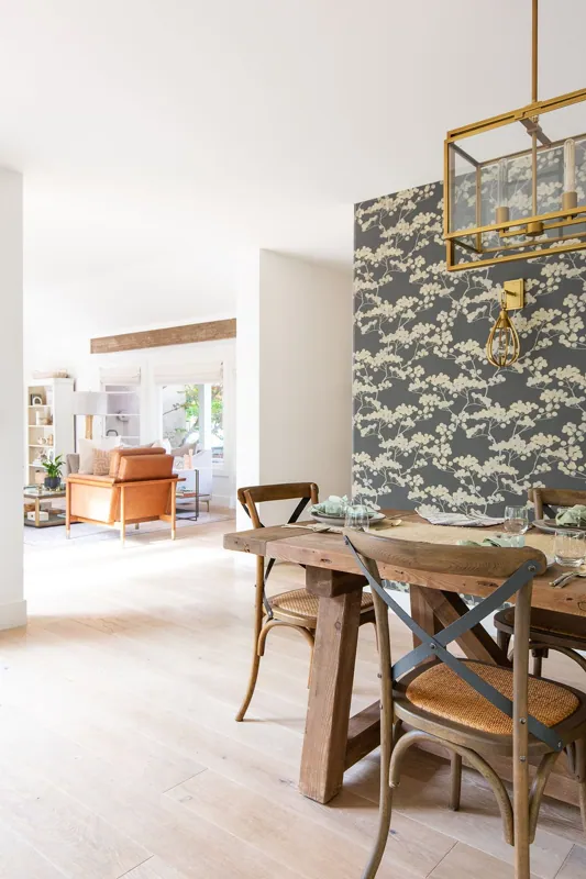 Part of a dining area with wooden table and chairs, and a decorated wallpaper on the wall. The background shows a living room with sofa and windows.