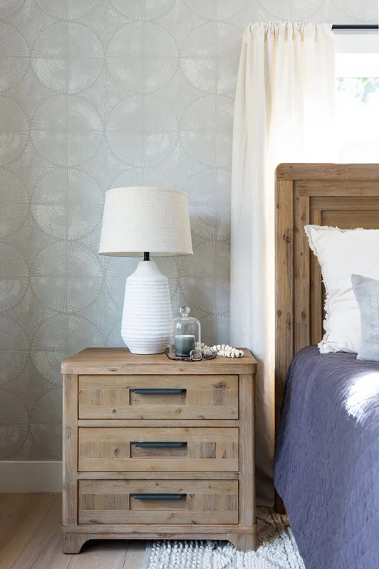 A wooden bedside table with three drawers, a white table lamp, a glass containing a candle and decorative shell, next to a bed with white and gray bedding, and a window with off-white curtains.