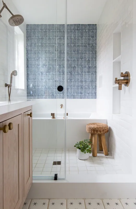 Bathroom with walk-in shower featuring patterned blue wall tiles, a wooden vanity with a white countertop and brass fixtures, a small woven stool, a potted plant, and a mix of white and patterned wall tiles.