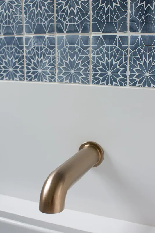 Close-up of a gold faucet handle mounted on a white wall below decorative blue and white mosaic tiles.