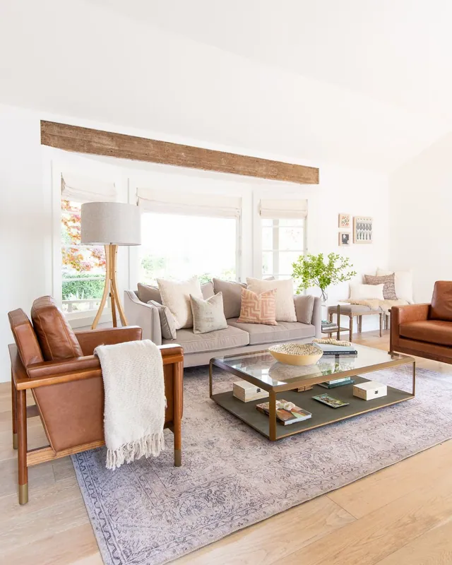 Bright living room with a beige sofa, leather armchair, coffee table, and decorative pillows, with large windows and wooden accents.