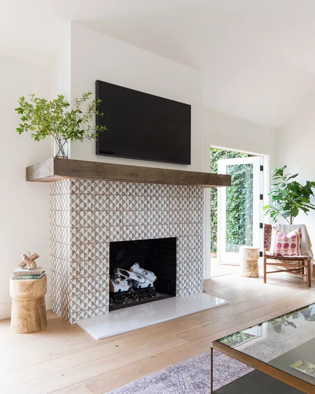 Living room with a modern mosaic fireplace, a mounted television above the fireplace, open door leading outside, green plants, a wooden chair with pillows, a small side table, and a glass coffee table.