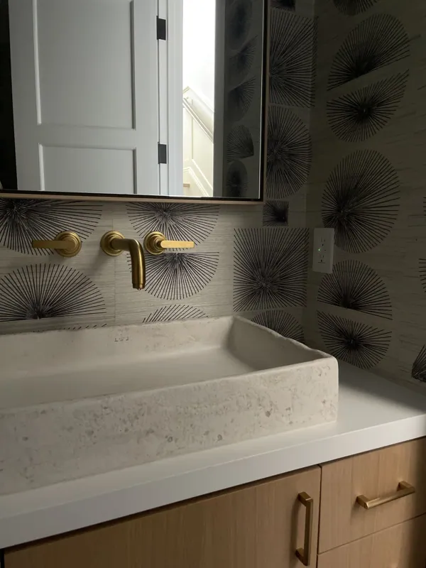 Bathroom vanity with a white stone sink, gold faucet and handles, wooden cabinet, a mirror, patterned wallpaper, and an electrical outlet.
