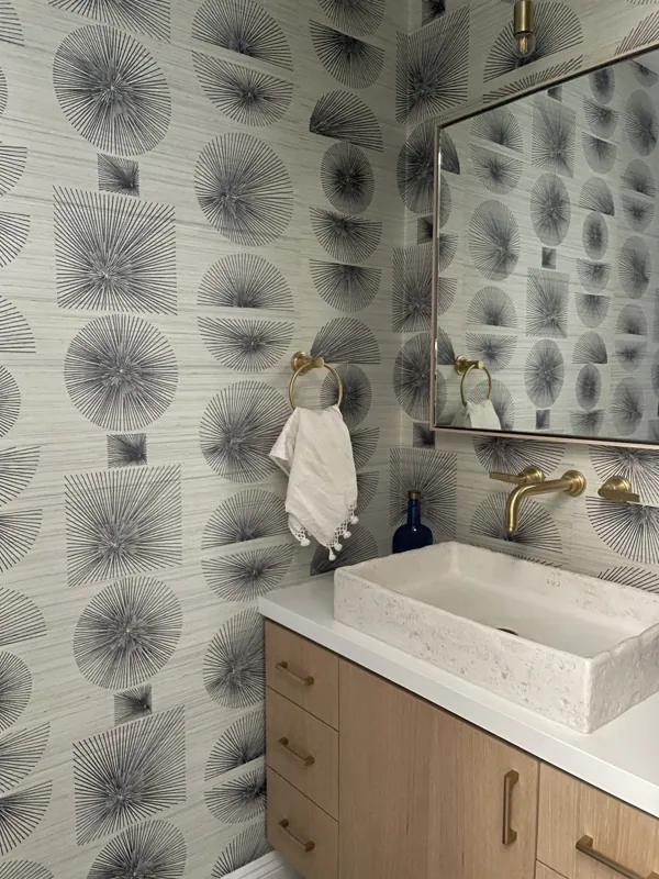 A powder room with patterned wallpaper in black geometric starburst designs, a large mirror, a wooden vanity with gold handles, a white rectangular stone sink, and a gold towel ring with a white towel and a blue decorative bottle.