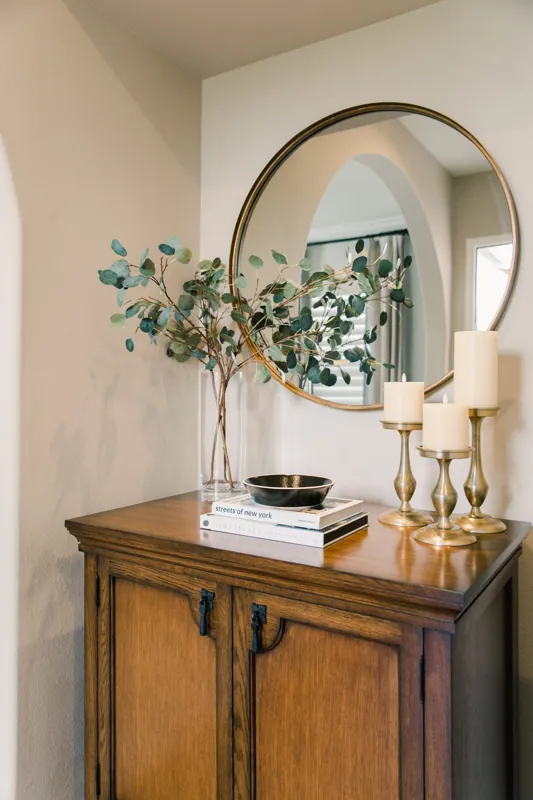 Decorative wooden cabinet with a mirror, a vase with greenery, three white candles on golden candlesticks, and a stack of books.
