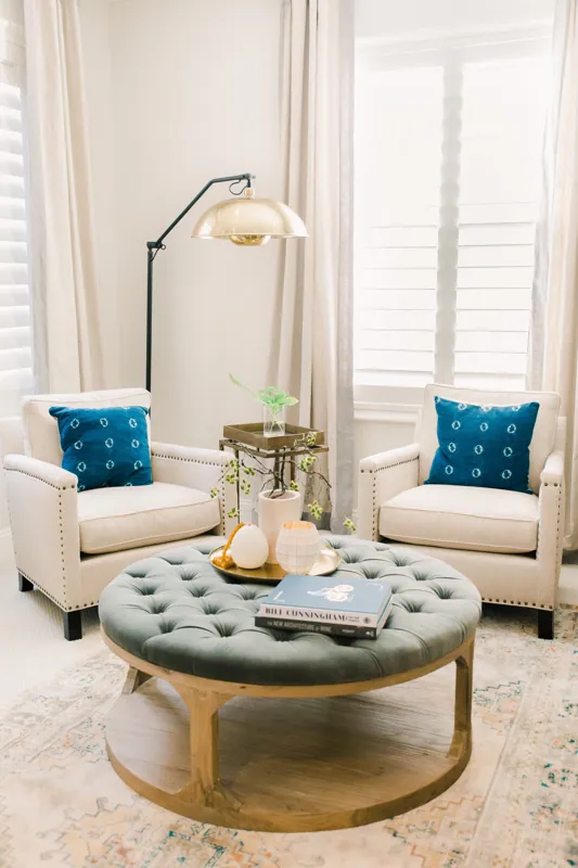 A bright living room with two cream armchairs decorated with blue pillows, a round tufted coffee table with books and decorative objects, and a large window with white shutters and beige curtains, illuminated by a gold-colored floor lamp.
