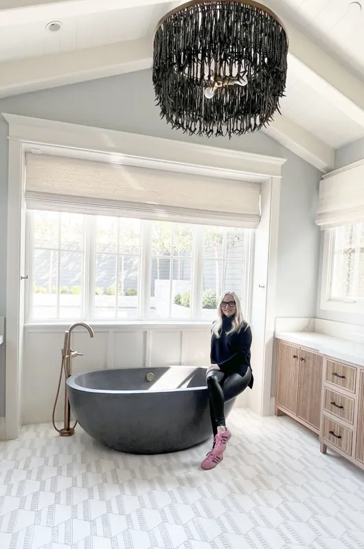 A woman sitting on the edge of a black bathtub in a bright bathroom with large windows, white walls, and a wooden cabinet.