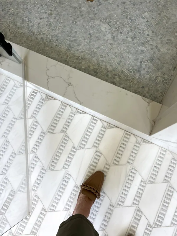 A person's foot wearing a tan loafer with a braided strap, stepping onto a white tiled floor next to a marble threshold and a glass shower door.