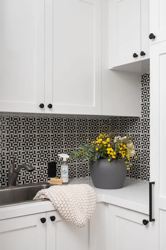 Kitchen sink area with white cabinets, black and white geometric backsplash, a gray plant pot with yellow flowers, hand soap spray, and a beige knitted towel.