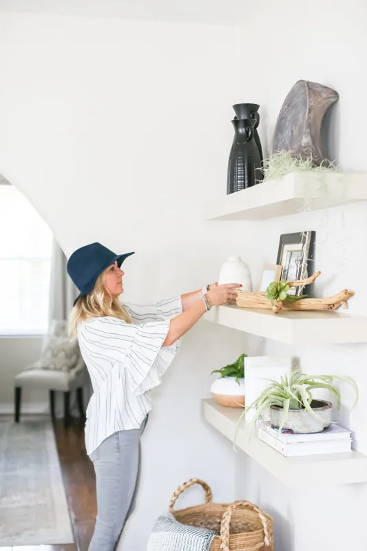 A woman in a striped top and blue hat arranged on a wall shelf with vases, picture frames, and decorative objects in a well-lit room.
