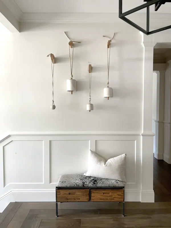 Modern interior with four hanging pendant lights above a white wainscoted wall, a bench with a patterned cushion and a white pillow, and hardwood floors.