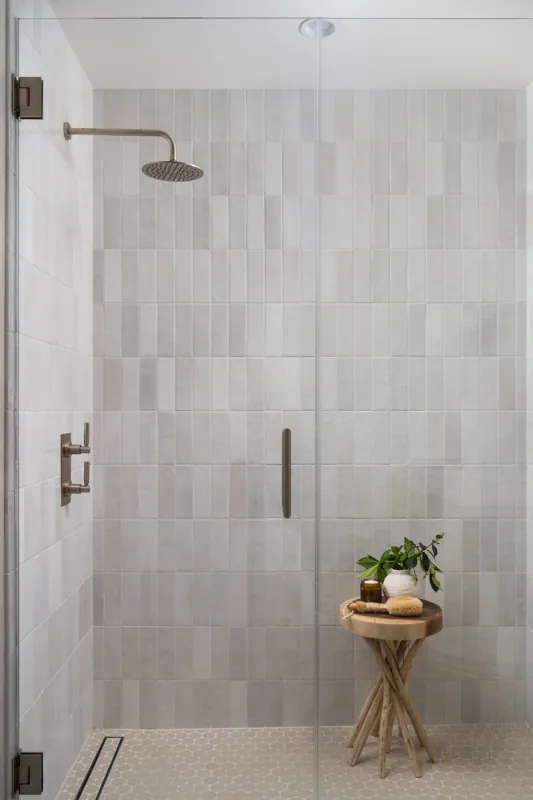 Modern glass shower enclosure with white tiled walls and a beige hexagonal tiled floor, a wooden stool with toiletries and a potted plant inside.