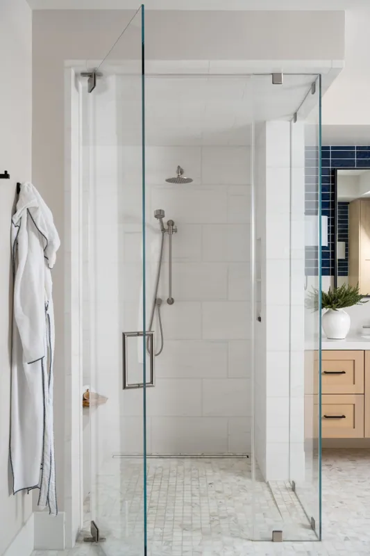 Walk-in glass shower with veined marble walls beside a wood vanity and a bright window.