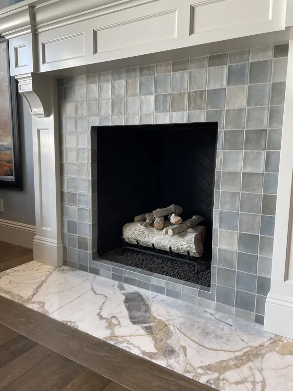 Bedroom fireplace clad in green zellige-style tile with a marble hearth and neatly stacked logs.