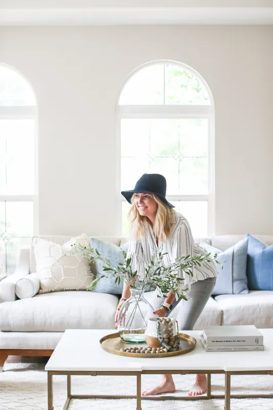 Designer in a hat styling a coffee table in a bright living room framed by tall arched windows.