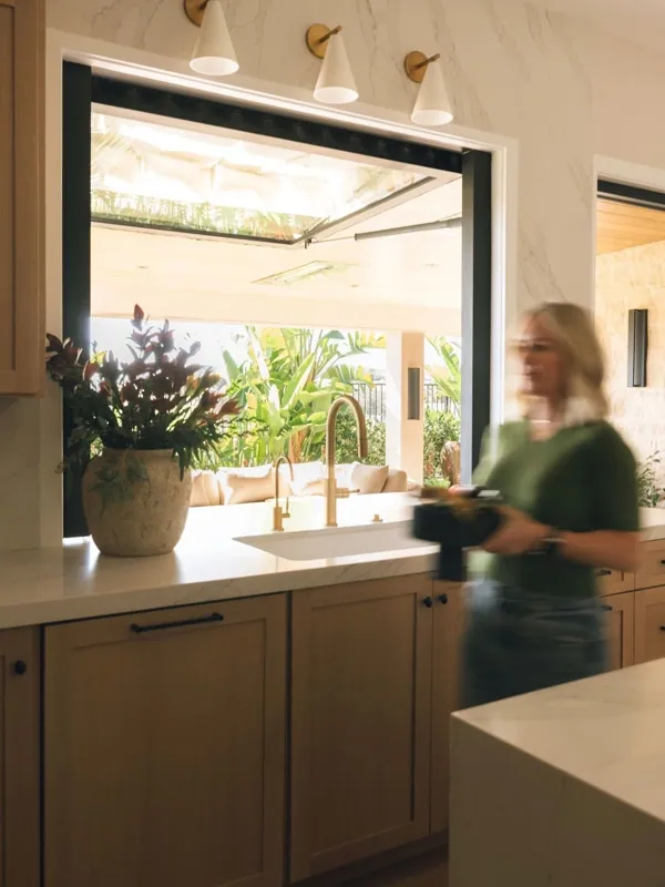 Meredith Gough at a kitchen sink with a pass-through window opening to a lush green patio, framed by warm wood cabinetry.