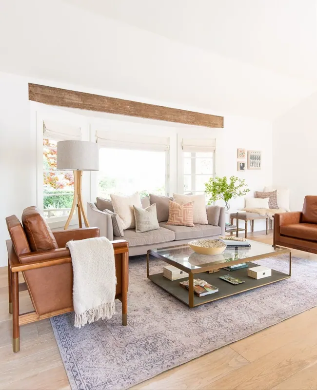 Bright, high-ceilinged living room with tan leather chairs, a linen sofa, a glass coffee table, and an exposed wood beam.