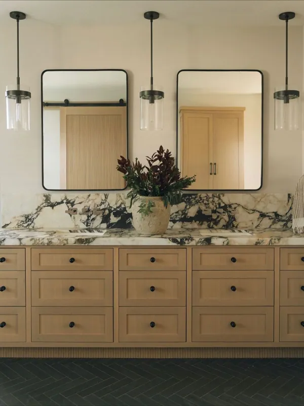 Double vanity with two wood cabinets, dramatic veined-marble counters, twin mirrors, and slim pendant lights.