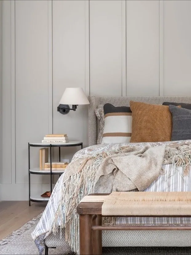 Quiet bedroom corner with layered neutral bedding, a wall-mounted sconce, paneled walls, and a wood bench.