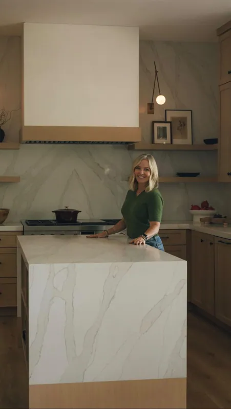 Meredith Gough behind a white marble waterfall island in a warm wood kitchen with a custom hood.