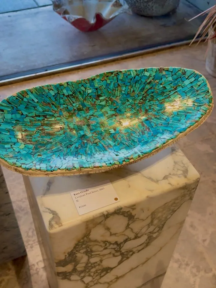 A hand-crafted teal glass art bowl displayed on a marble pedestal, part of a curated art selection.